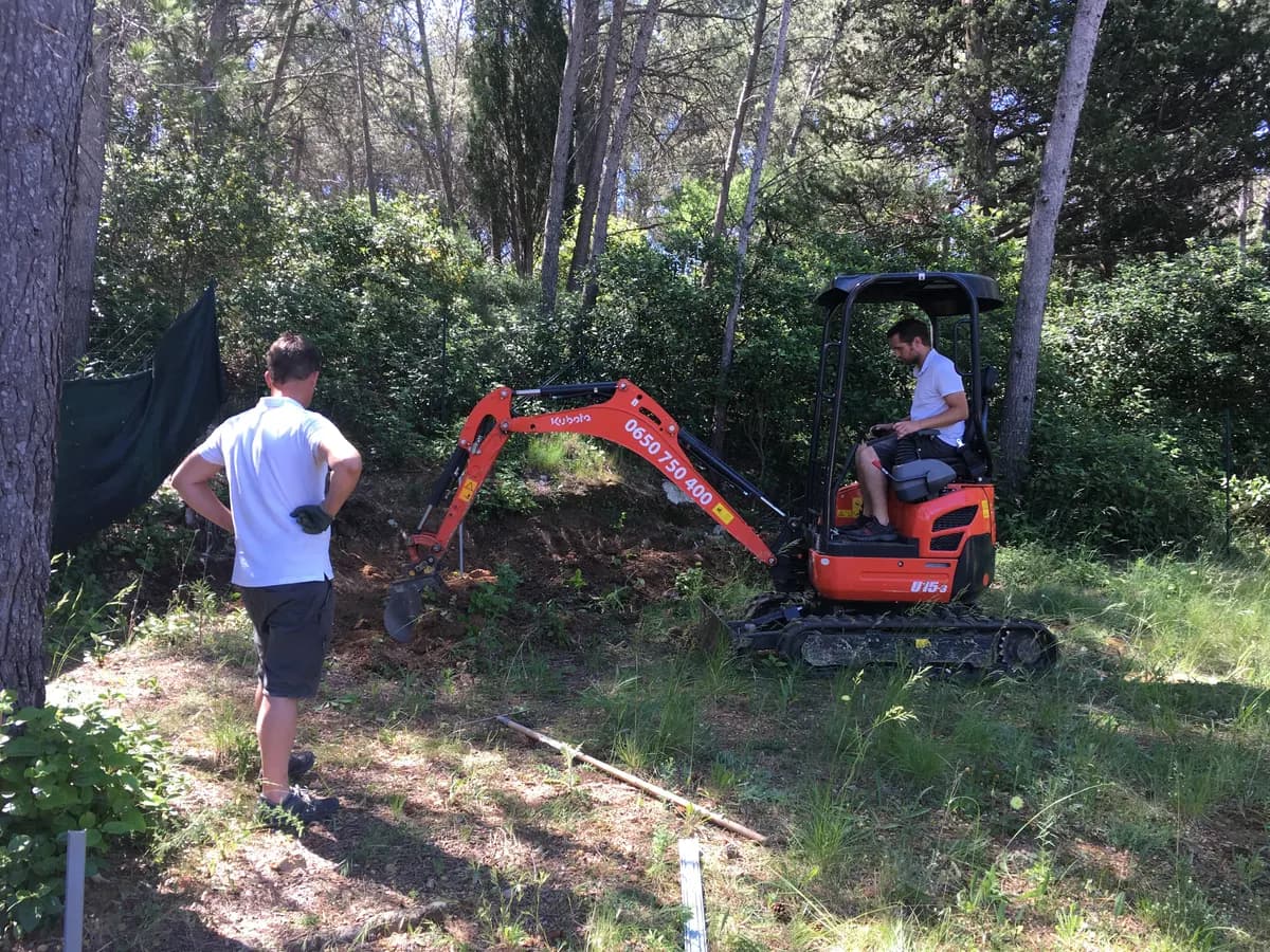 Mini-pelle Loc'Family attaquant un talus envahi de broussailles, sous les pins d'un jardin de Jacou