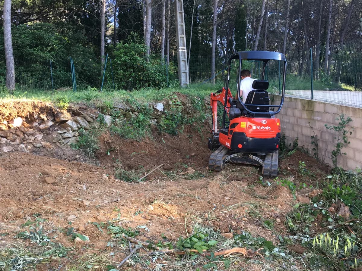 Talus déblayé au pied d'un ancien muret de pierres sèches, la mini-pelle calée contre le mur de clôture